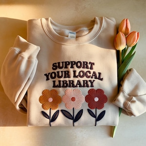 Puede incluir: Una sudadera beige con el texto bordado "SUPPORT YOUR LOCAL LIBRARY" en marrón. Debajo, tres flores bordadas en naranja, rosa y rojo. Un ramo de tulipanes descansa junto a la sudadera.