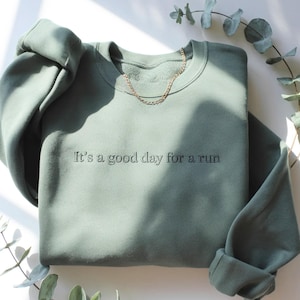 May include: Sage green sweatshirt with the embroidered text "It's a good day for a run." A gold chain necklace rests on the crew neck. The sleeves are folded up. Eucalyptus leaves are in the background.