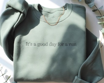 Puede incluir: Una sudadera de cuello redondo en color verde salvia con el texto bordado "It's a good day for a run" en hilo verde oscuro a juego en el pecho. La prenda tiene mangas largas con puños acanalados y un cuello acanalado de diseño sencillo.