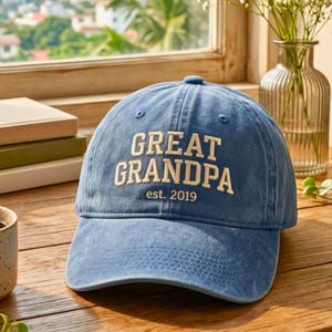 May include: A blue baseball cap with the words "GREAT GRANDPA" embroidered in white, with "est. 2019" below. The hat is sitting on a wooden surface near a window and a vase of flowers.