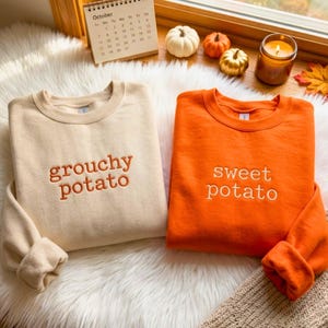 May include: A cream-colored sweatshirt with the embroidered text "grouchy potato" and an orange sweatshirt with the embroidered text "sweet potato". Both feature crew necklines and long sleeves, displayed on a white faux fur surface.