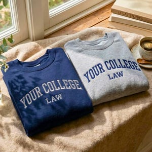 May include: Two crewneck sweatshirts, one navy blue and one heather gray, are displayed. Both feature the embroidered text "YOUR COLLEGE LAW" in white and blue, respectively. The sweatshirts are folded and rest on a beige blanket.