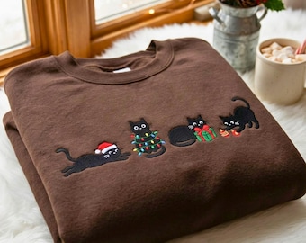 May include: A brown crewneck sweatshirt featuring an embroidered design of four black cats. One cat wears a Santa hat, one is tangled in colorful Christmas lights, one sits with a green gift box, and one plays with red and gold ornaments.