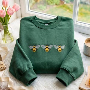 May include: A forest green crewneck sweatshirt featuring an embroidered design of three honeybees across the center chest. Each bee is stitched with yellow and black horizontal stripes and white wings. The garment has long sleeves with ribbed cuffs.