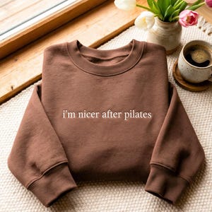 Puede incluir: Una sudadera marrón de cuello redondo con el texto bordado en blanco "i'm nicer after pilates" centrado en el pecho. La prenda tiene mangas largas con puños acanalados y un cuello acanalado, confeccionada en una tela de color sólido.