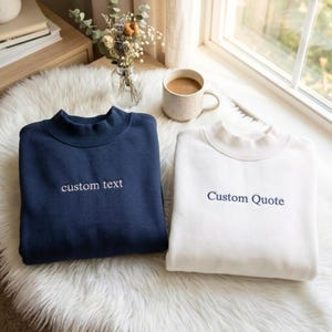 May include: A navy blue sweatshirt with the embroidered text custom text and a white sweatshirt with the embroidered text Custom Quote. Both garments feature a mock neck collar and long sleeves, presented side by side on a soft white surface.