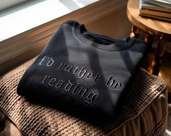 May include: Black sweatshirt with the embroidered phrase "I'd rather be reading." The crew neck sweatshirt is folded on a brown, textured ottoman. A small wooden side table and books are in the background.