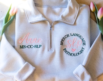 May include: A light gray quarter-zip sweatshirt featuring embroidered text. The left side displays the name Anne in pink and MS-CC-SLP in dark green. The right side features a circular design with Speech Language Pathologist and SLP in pink.