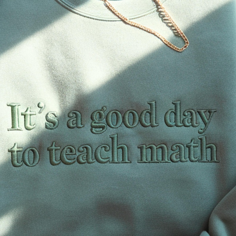 Puede incluir: Sudadera verde claro con la frase bordada "It's a good day to teach math." Una cadena dorada est&aacute; colocada en la parte superior. La sudadera es de color liso y tiene cuello redondo.