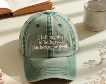 May include: A sage green baseball cap with the embroidered text "I left my dog to be here.... This better be good." The hat has a curved brim and a classic six-panel design. The hat is sitting on a light green surface.