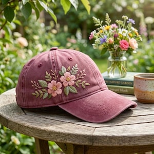 May include: A maroon baseball cap with embroidered pink flowers and green leaves. The cap is sitting on a wooden table outdoors, with a blurred background of greenery and flowers.
