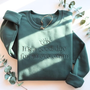 Könnte beinhalten: Ein teal-grünes Sweatshirt mit dem Schriftzug "It's a good day for an eye exam" und einer Augen-Grafik. Eine goldene Halskette liegt auf dem gefalteten Sweatshirt. Die Ärmel sind hochgekrempelt.