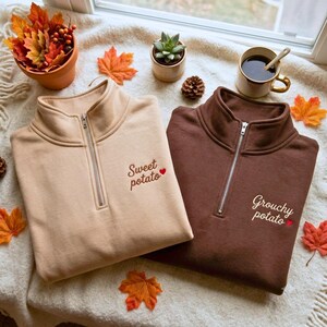 May include: Two quarter-zip sweatshirts, one in beige and one in dark brown, each featuring embroidered text on the left chest. The beige sweatshirt reads Sweet potato with a small red heart, and the brown sweatshirt reads Grouchy potato with a red heart.