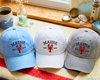 May include: Three baseball caps in light blue, white, and grey. Each cap features embroidered text that reads MAINE EST. 1820 above a red lobster graphic. The caps are made of a soft fabric and have a structured crown with a curved brim.
