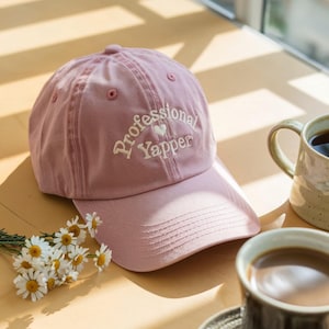 May include: A dusty rose baseball cap with the embroidered text "Professional Yapper" in white script, featuring a small heart. The cap is on a light-colored surface, with a cup of coffee and flowers in the background.