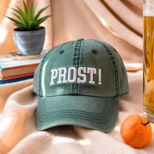 May include: A green baseball cap with the word "PROST!" embroidered in white. The cap is sitting on a beige surface, with a small orange fruit and a glass of beer in the background. A small potted plant and books are also visible.