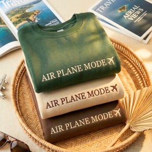 Puede incluir: Tres sudaderas plegadas en verde, beige y marr&oacute;n, apiladas en una bandeja tejida. Cada sudadera tiene el texto "AIR PLANE MODE" con un avi&oacute;n. Revistas de viajes y gafas de sol en el fondo.