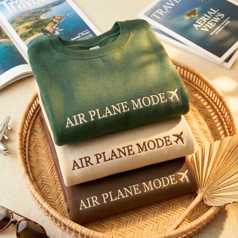 Puede incluir: Tres sudaderas plegadas en verde, beige y marr&oacute;n, apiladas en una bandeja tejida. Cada sudadera tiene el texto "AIR PLANE MODE" con un avi&oacute;n. Revistas de viajes y gafas de sol en el fondo.