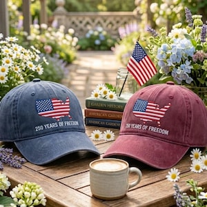 May include: A blue baseball cap and a maroon baseball cap, each featuring an embroidered map of the United States filled with the American flag pattern and the text 250 YEARS OF FREEDOM. Both caps are positioned on a wooden surface near a coffee mug.