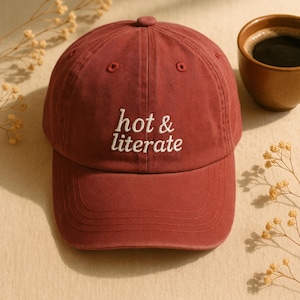 May include: A maroon baseball cap with the embroidered text "hot & literate" in white. The cap has a curved brim and a button on top. The image also includes a cup of coffee and decorative sprigs.