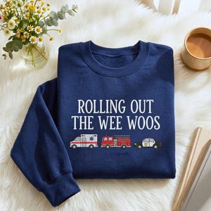 Puede incluir: Sudadera azul marino con el texto blanco "ROLLING OUT THE WEE WOOS". Debajo del texto, ilustraciones de una ambulancia, un camión de bomberos y un coche de policía.