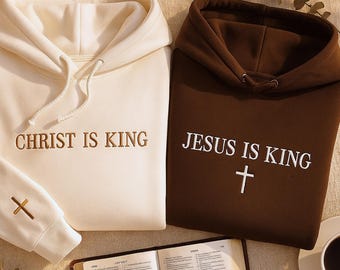 May include: Two hooded sweatshirts, one cream-colored and one brown, with the text "CHRIST IS KING" and "JESUS IS KING" embroidered on them. A cross is embroidered on the cream-colored sweatshirt's sleeve. A Bible and a cup of coffee are also in the image.