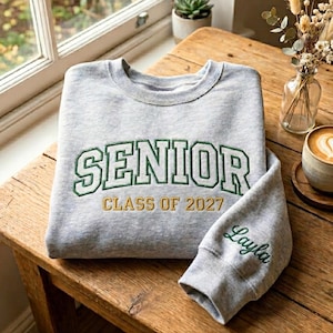 May include: A gray sweatshirt with the word "SENIOR" in green and white lettering, with "CLASS OF 2027" in gold lettering. The sleeve has the name "Layla" embroidered in green script.