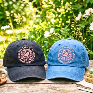 May include: Two baseball caps, one in charcoal gray and one in light blue, each featuring a circular embroidered design in pink. The design depicts a bird surrounded by flowers and leaves, with the text "RADICALIZED BY BASIC DECENCY" encircling the central illustration.