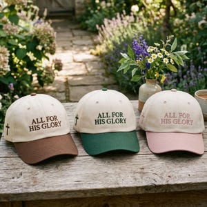 May include: Three baseball caps with cream-colored crowns and colored brims in brown, dark green, and light pink. Each cap features the embroidered text ALL FOR HIS GLORY on the front and a small cross symbol on the side panel in matching thread.