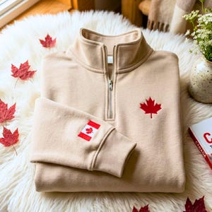 May include: A beige quarter-zip sweatshirt featuring a red embroidered maple leaf on the chest and a small Canadian flag patch on the sleeve. The garment has a high collar with a silver-toned metal zipper and ribbed cuffs on the long sleeves.