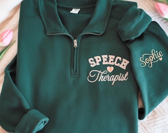 May include: A dark green quarter-zip sweatshirt featuring light pink embroidery on the chest that reads "SPEECH Therapist" with a small heart icon. The left sleeve is embroidered with the name "Sophie" in a cursive font and a small heart.