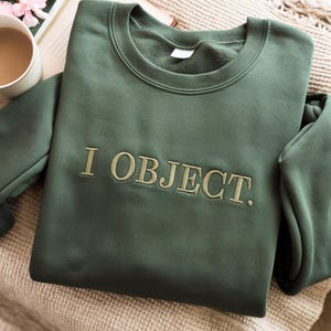 Puede incluir: Una sudadera verde oscuro con el texto bordado "I OBJECT.". La sudadera de cuello redondo está hecha de un material suave. El texto es de un tono verde más claro.