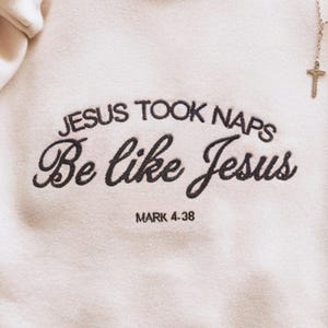 May include: A cream-coloured sweatshirt featuring the embroidered phrase "JESUS TOOK NAPS Be like Jesus" in black script, with "MARK 4-38" below. A gold cross pendant is visible in the top right corner.