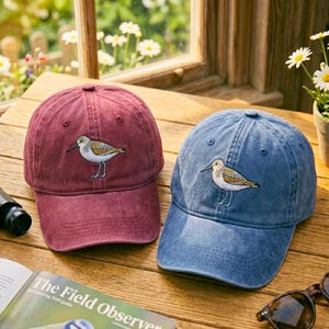 Puede incluir: Dos gorras de béisbol, una burdeos y otra azul, cada una bordada con un diseño de pájaro. Las gorras están sobre una superficie de madera junto a una guía de campo y gafas de sol. Las gorras son de estilo clásico e informal.