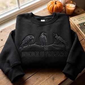 Puede incluir: Una sudadera negra de cuello redondo con un diseño bordado de tres cuervos posados en una rama. Debajo de las aves, el texto "MURDER IN PROGRESS" aparece en una fuente de estilo gótico. El bordado está realizado en tonos grises y blancos.