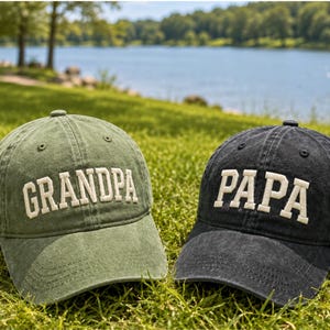 May include: Two baseball caps with embroidered text. One is olive green with "GRANDPA" in white block letters. The other is black with "PAPA" in white block letters. Both caps are on a grassy surface with a lake and trees in the background.