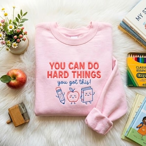 May include: A light pink crewneck sweatshirt featuring the embroidered text "YOU CAN DO HARD THINGS you got this!" in red and blue. Below the text are three small, smiling cartoon characters: a pencil, an apple, and a closed book.