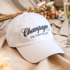 May include: A white baseball cap with the word "Champagne" embroidered in black script, along with "S'IL VOUS PLAÎT" in a smaller font. The cap has a curved brim and a button on top. The background includes a champagne glass and dried flowers.