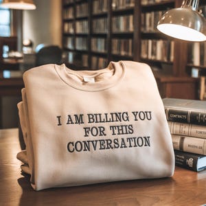 May include: Beige sweatshirt with black embroidered text reading "I AM BILLING YOU FOR THIS CONVERSATION". The sweatshirt is stacked with others on a wooden table. Books and a lamp are in the background, suggesting a library setting.