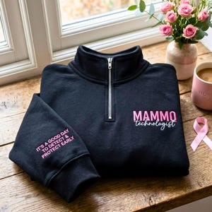 May include: A black quarter-zip sweatshirt featuring pink embroidered text on the chest that reads MAMMO technologist and on the sleeve that reads IT'S A GOOD DAY TO DETECT & PROTECT EARLY. The garment has a silver zipper and long sleeves.