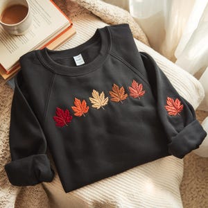 May include: A black sweatshirt with embroidered fall leaves in shades of red, orange, and gold. The leaves are arranged across the chest and on the left sleeve. The sweatshirt is folded on a cream-colored blanket.