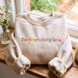May include: A beige crewneck sweatshirt featuring the embroidered text "i'm nicer after pilates" in multicolored letters. The design includes small embroidered star shapes in various colors surrounding the text on the front of the garment.