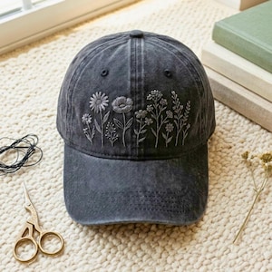 May include: A charcoal gray baseball cap featuring detailed white embroidered wildflowers across the front panels. The cap has a distressed, washed-out fabric finish, a curved brim, and a structured crown with visible stitching along the seams.
