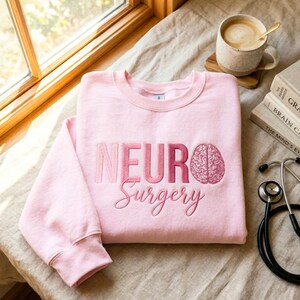 Puede incluir: Sudadera rosa claro con las palabras "NEURO Surgery" bordadas en tonos rosas y un dibujo de un cerebro. Un estetoscopio, libros y una taza de café son visibles en el fondo.