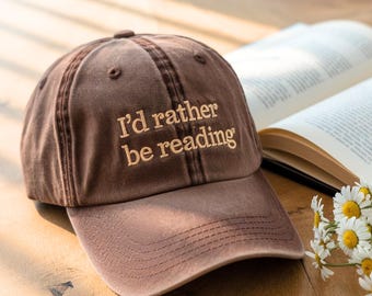 May include: A brown baseball cap with the embroidered text "I'd rather be reading" in a lighter brown color. The cap is sitting on a wooden surface next to an open book and a small bouquet of white flowers.