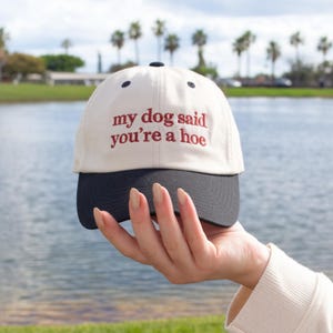 May include: A baseball cap with a cream-colored crown and a black brim. The cap features the embroidered phrase "my dog said you're a hoe" in red lettering. The cap is held up against a backdrop of a lake and palm trees.