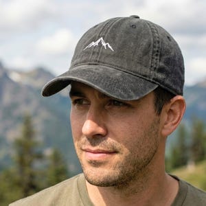 May include: A charcoal gray baseball cap with a white mountain range embroidered on the front. The cap has a curved brim and a distressed, vintage look. The background features a mountain range.