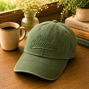 May include: A green baseball cap with the embroidered text "Great Grandpa". The cap has a curved brim and a classic six-panel design. The hat is made of a soft, worn fabric.