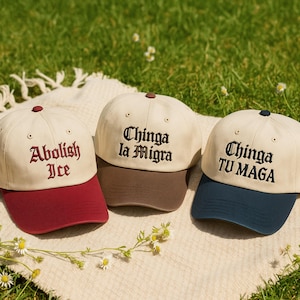May include: Three baseball caps with different color schemes and embroidered text. The caps have a cream-colored crown and a colored brim. One cap has "Abolish Ice" in burgundy, another "Chinga la Migra" in brown, and the last "Chinga TU MAGA" in navy.