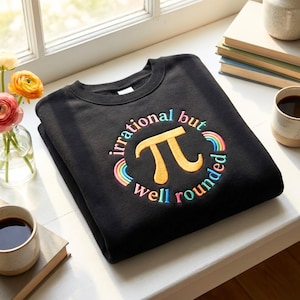 以下が含まれることがあります： 黒色のスウェットシャツ。刺繍された「irrational but well rounded」の文字が、金色のπ（パイ）のシンボルを囲んでいます。テキストとシンボルは虹のデザインで囲まれています。スウェットシャツは白い表面に折りたたまれています。